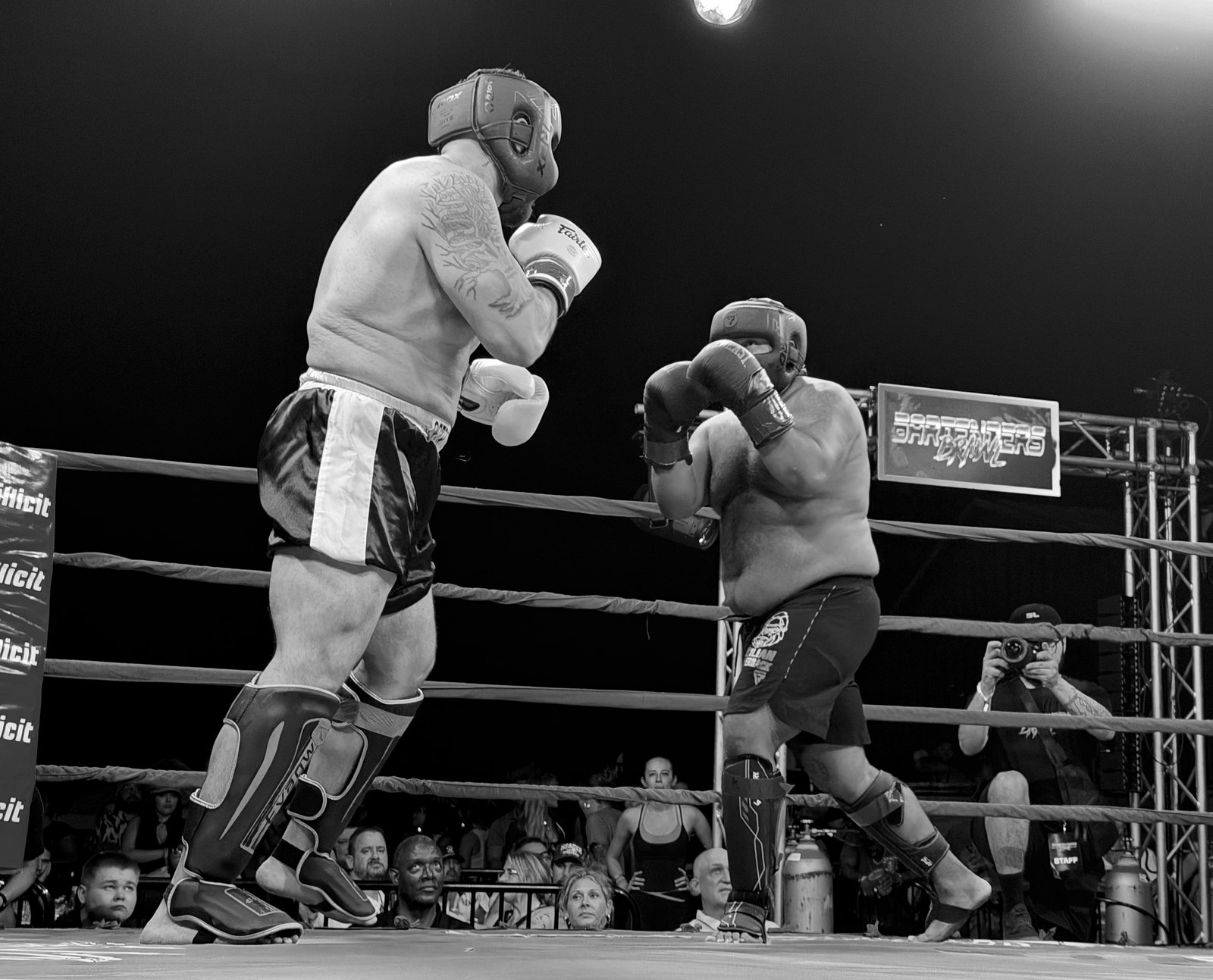 Two heavyweight fighters in headgear and shin guards square off in the ring, with the crowd and ringside photographers behind the ropes.