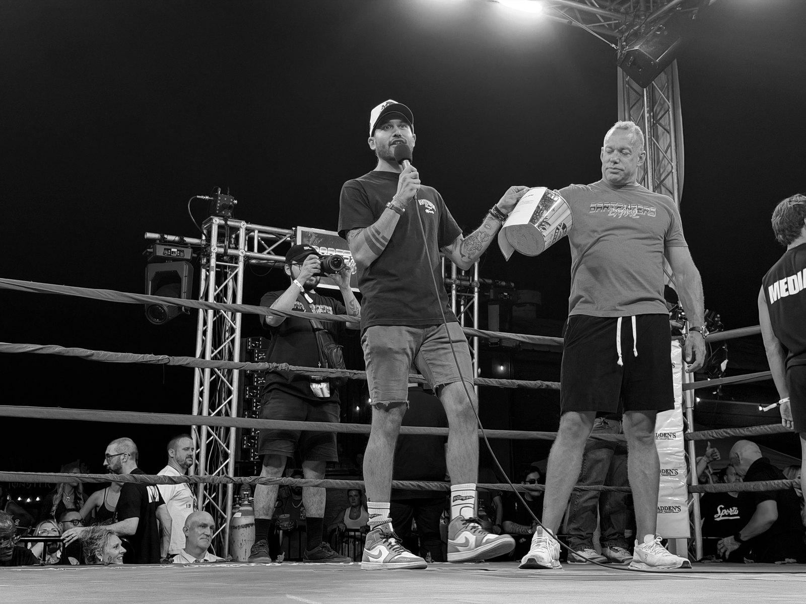 A man in a ball cap speaks into a microphone at the center of the ring while another man in a Bartenders Brawl shirt holds the raffle bucket beside him.