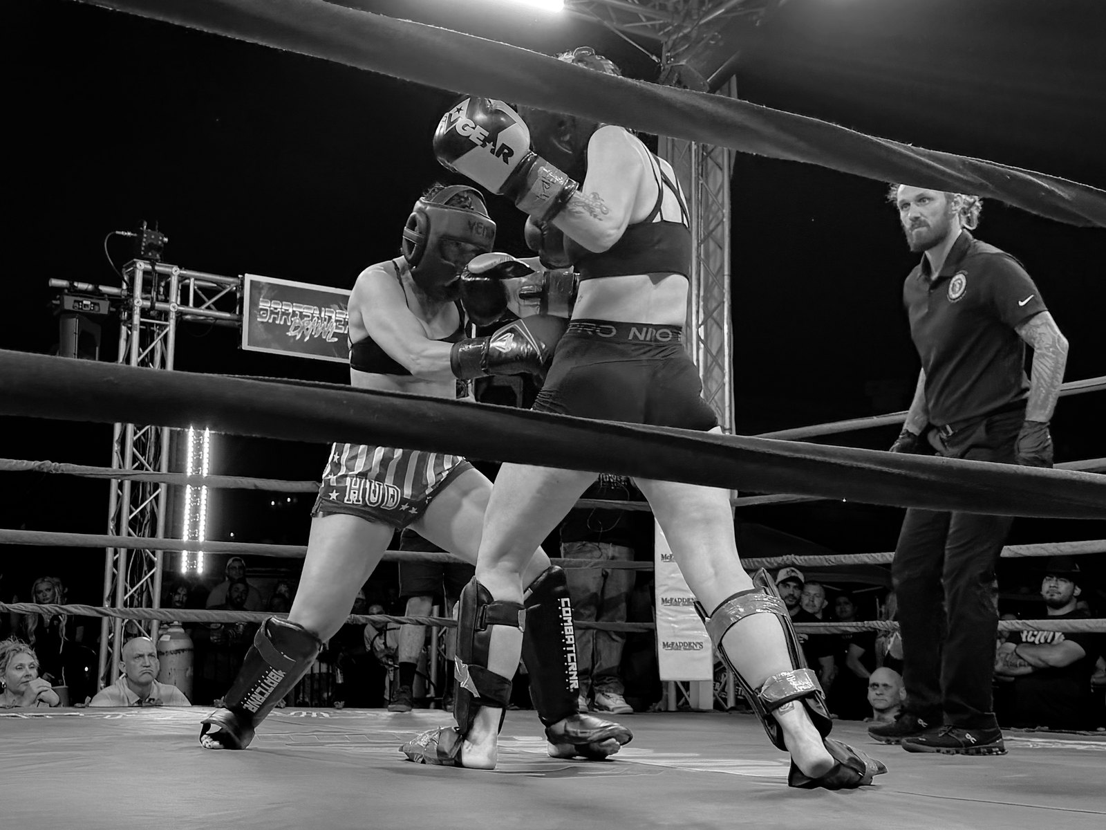 Two fighters trade at close range in the ring as the referee watches, the crowd pressed against the ropes beyond.